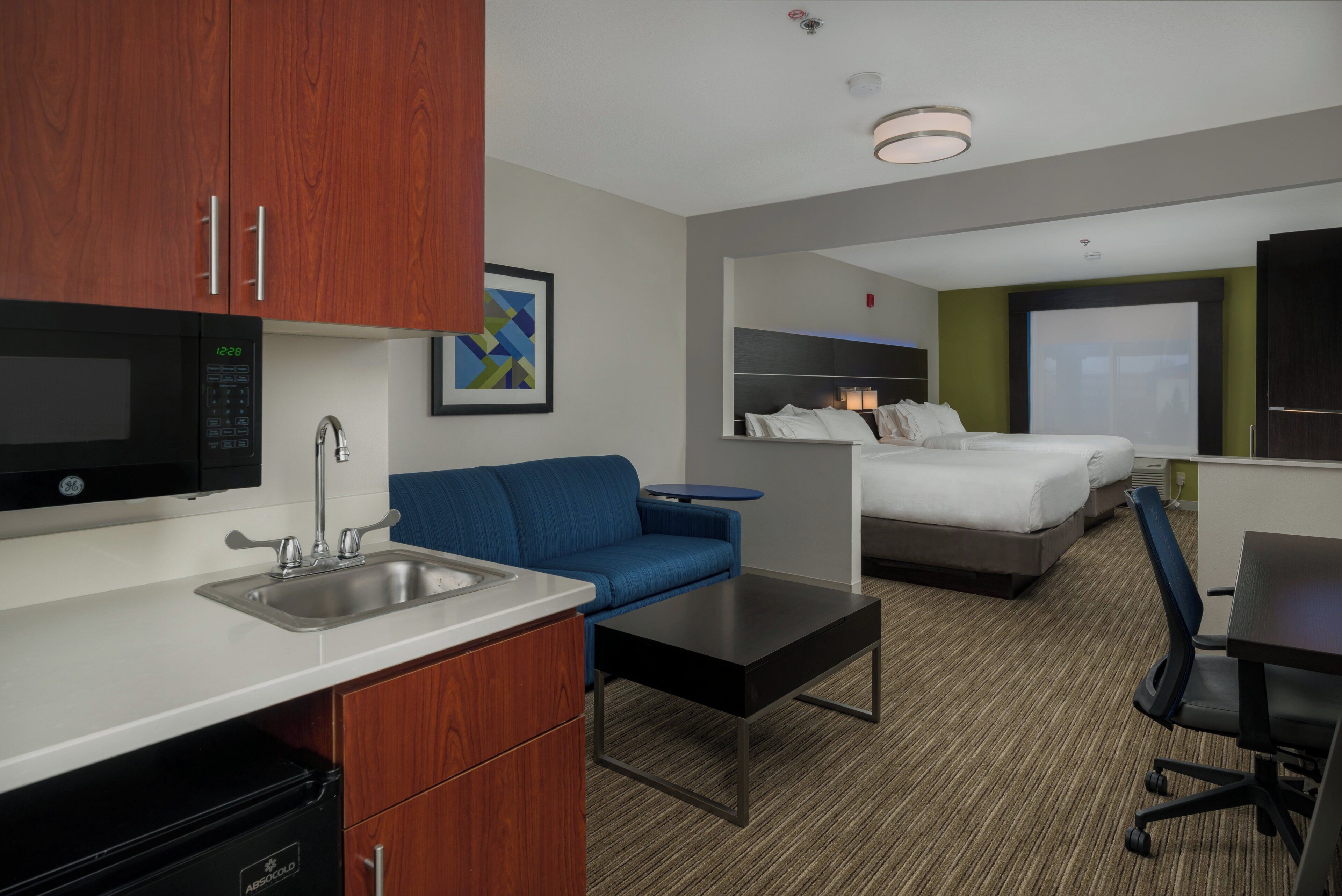 A hotel room with a kitchenette, a bed, a blue sofa, a desk, and a large window.