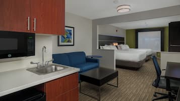 A hotel room with a kitchenette, a bed, a blue sofa, a desk, and a large window.