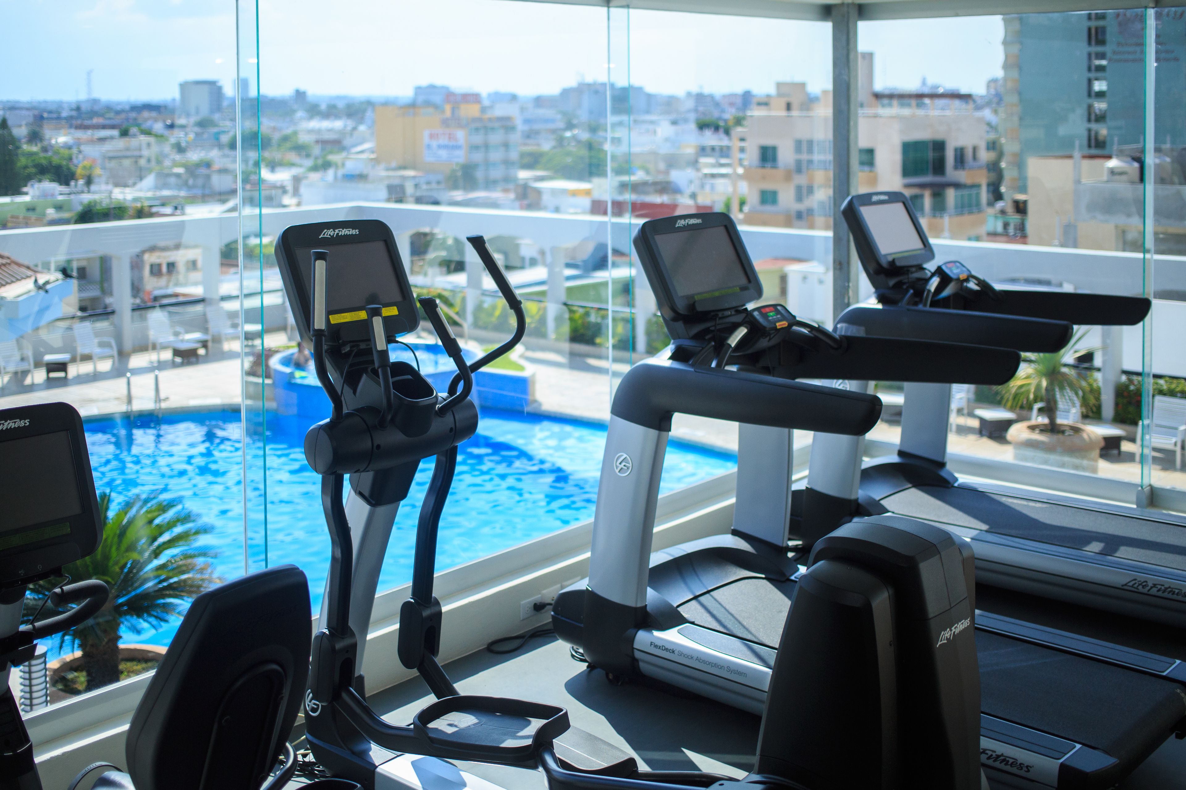 Ein Fitnessstudio mit Trainingsgeräten und Blick auf ein Schwimmbad und die Skyline der Stadt.