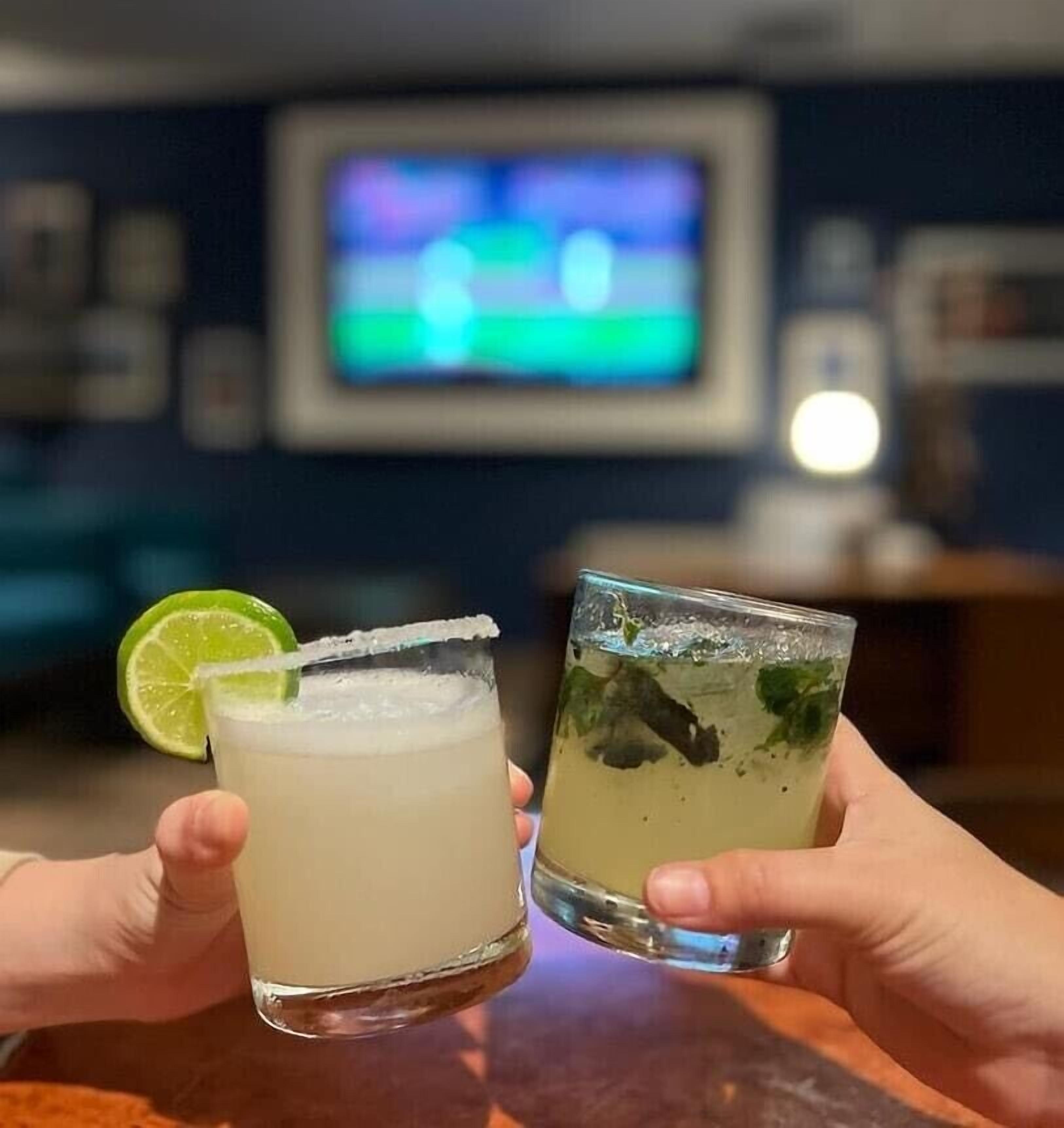 Dos bebidas con adorno de lima y menta, sujetas por manos, con un televisor al fondo.