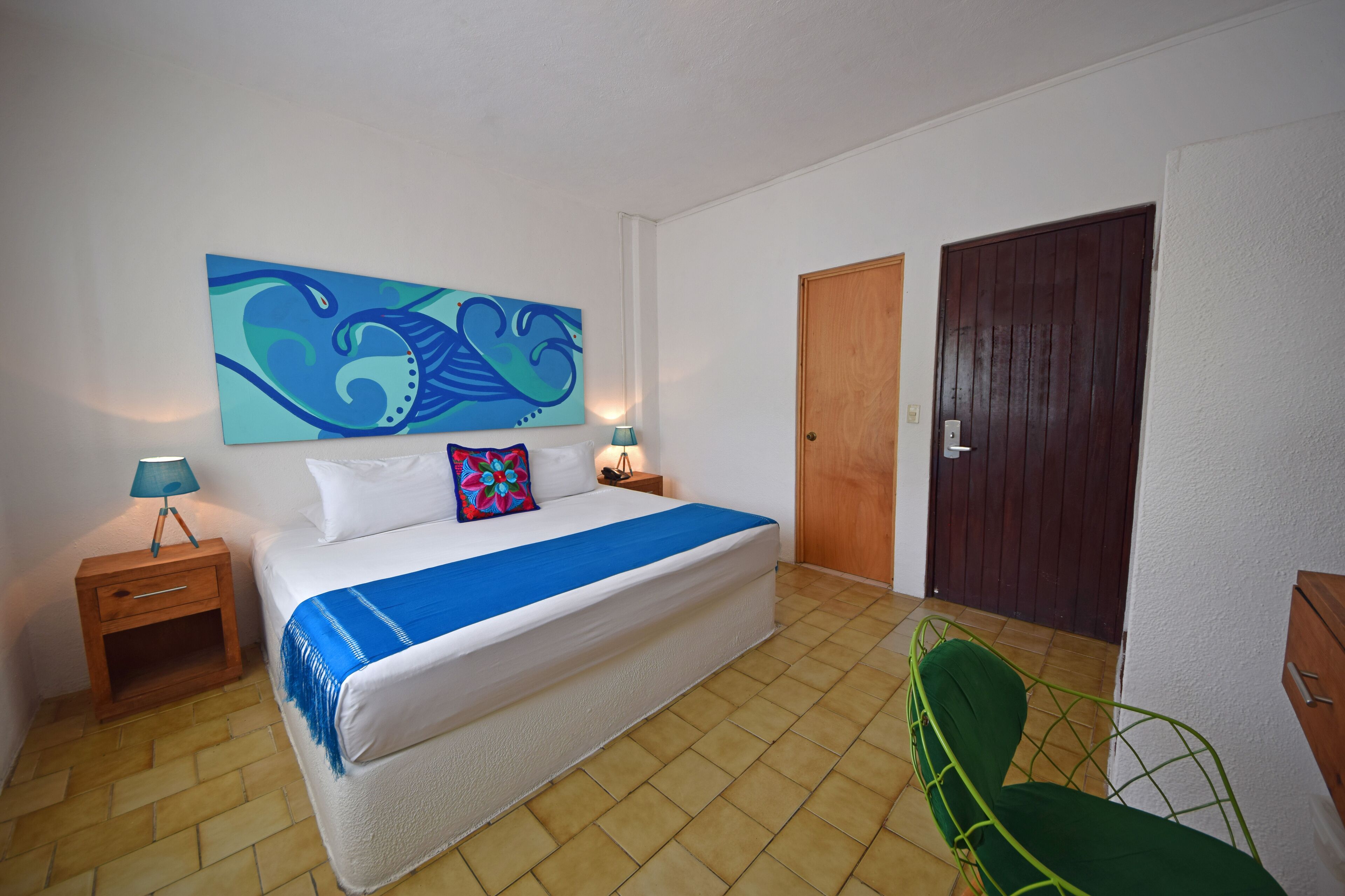 A bedroom with a bed, a nightstand, a lamp, a green chair, a wooden door, and a wall painting.