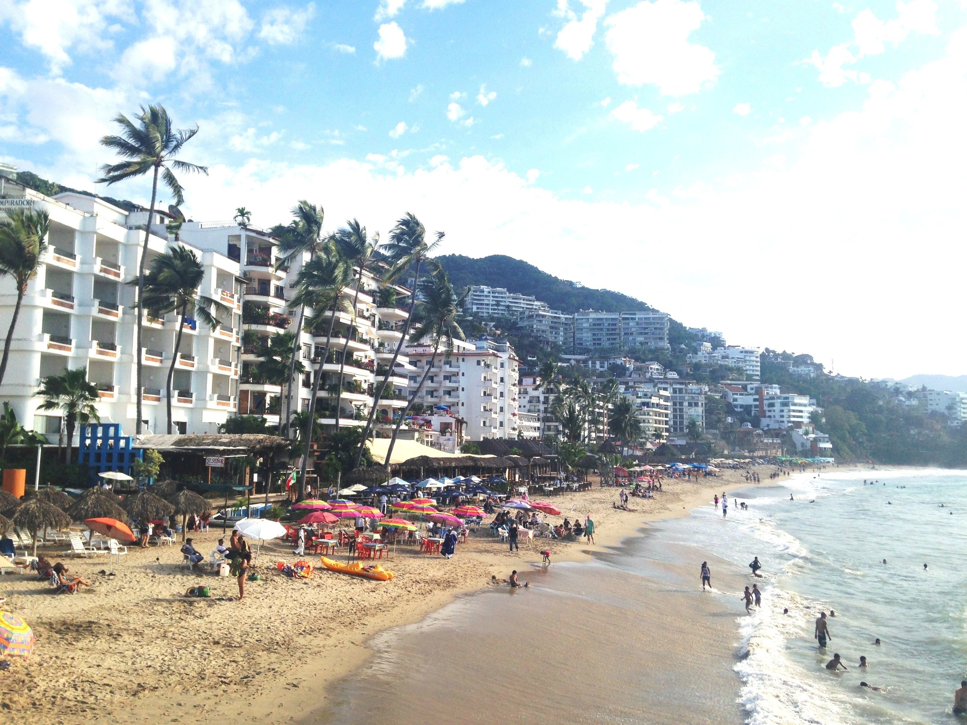 Una spiaggia con edifici a più piani, palme e ombrelloni.