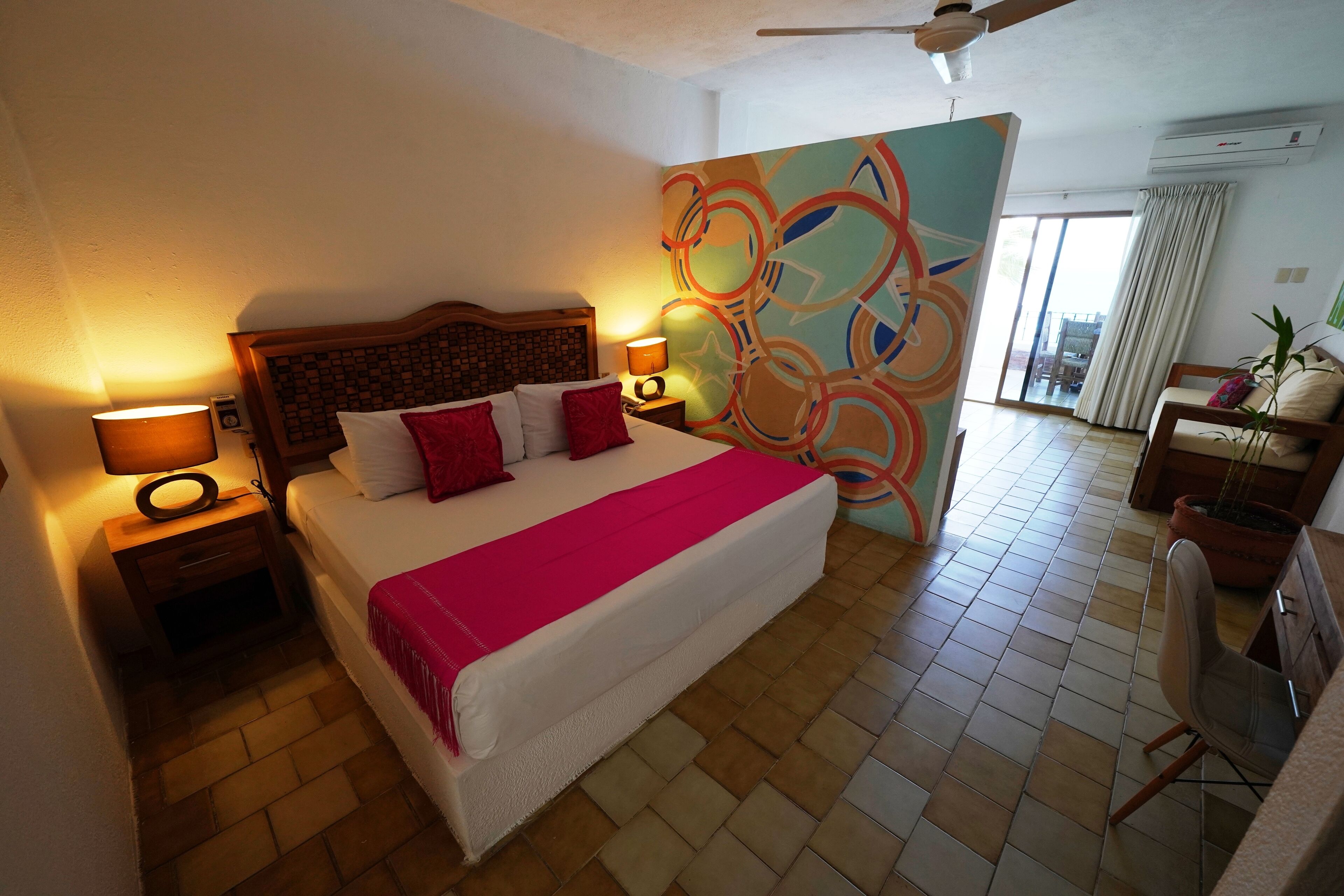Habitación de hotel con una cama grande, dos mesitas de noche con lámparas, un ventilador de techo, una pared con un diseño abstracto colorido y vistas al exterior a través de una puerta corredera.