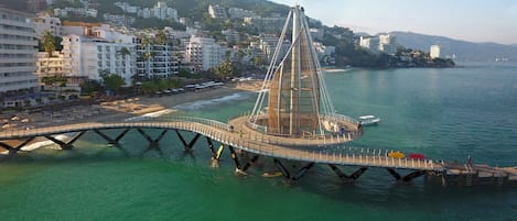 A cable-stayed bridge with a central tower, connecting a coastal area with a hillside residential area.