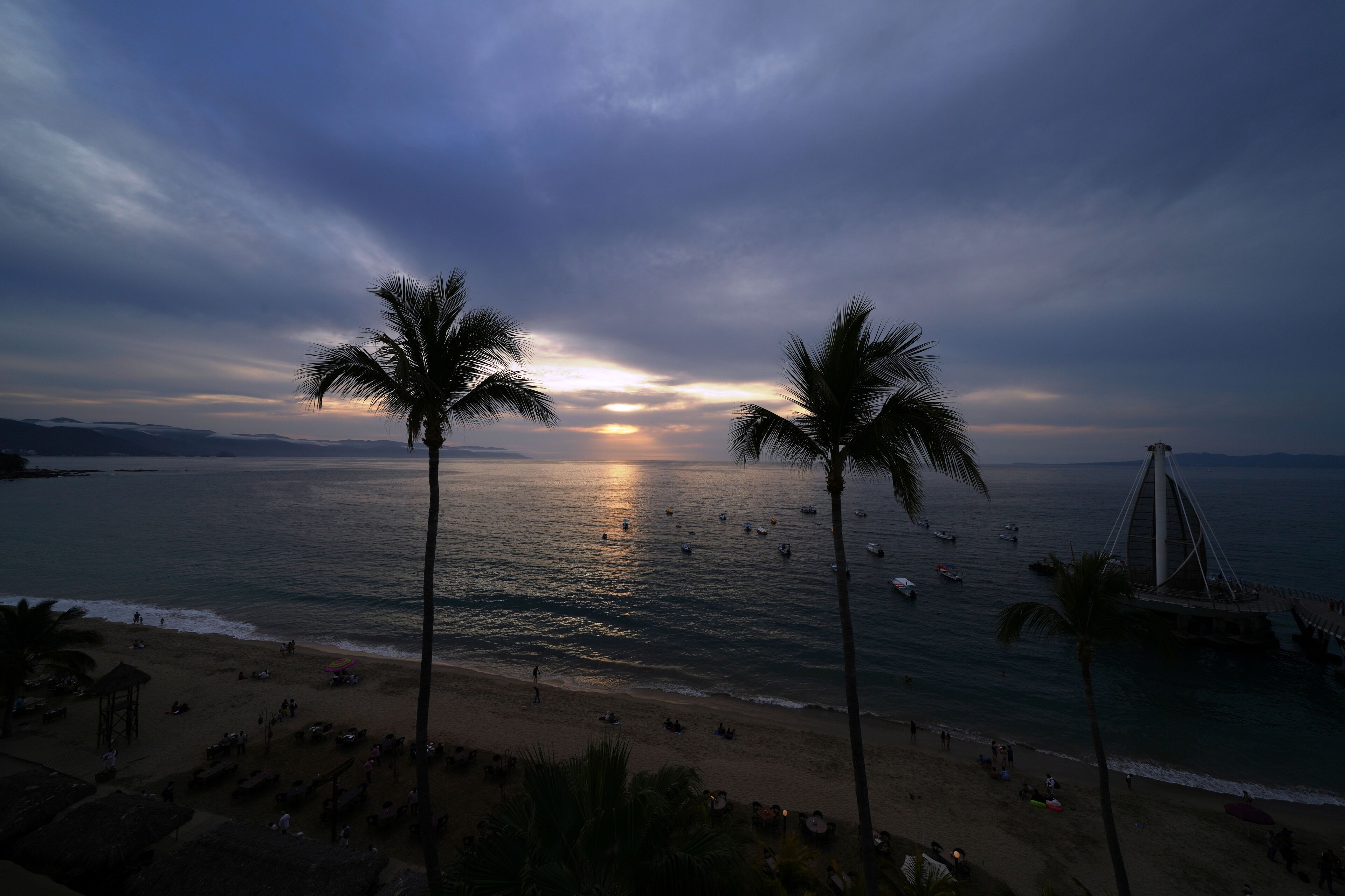 A beach with palm trees, a bridge, and boats on the water.