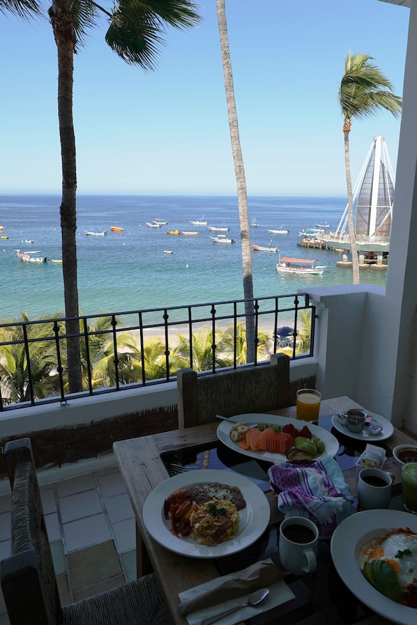 A balcony with a view of the ocean, palm trees, and a bridge. There are plates of food and drinks on the table. - Emperador Vallarta Beachfront Hotel and Suites (Puerto Vallarta)