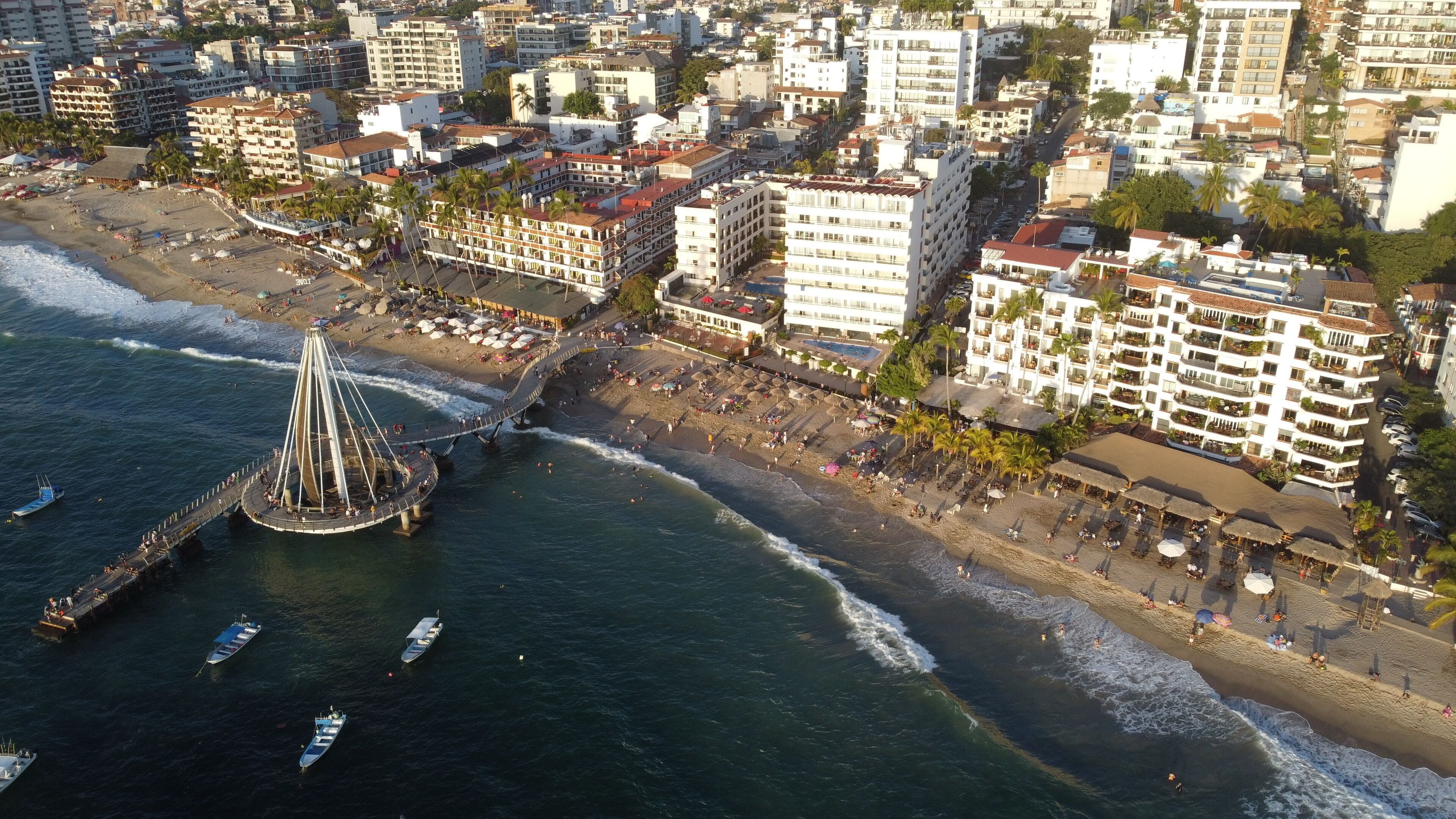 Vue aérienne d’une ville côtière, avec une tour imposante, des hôtels en bord de mer et une jetée où sont amarrés des bateaux.