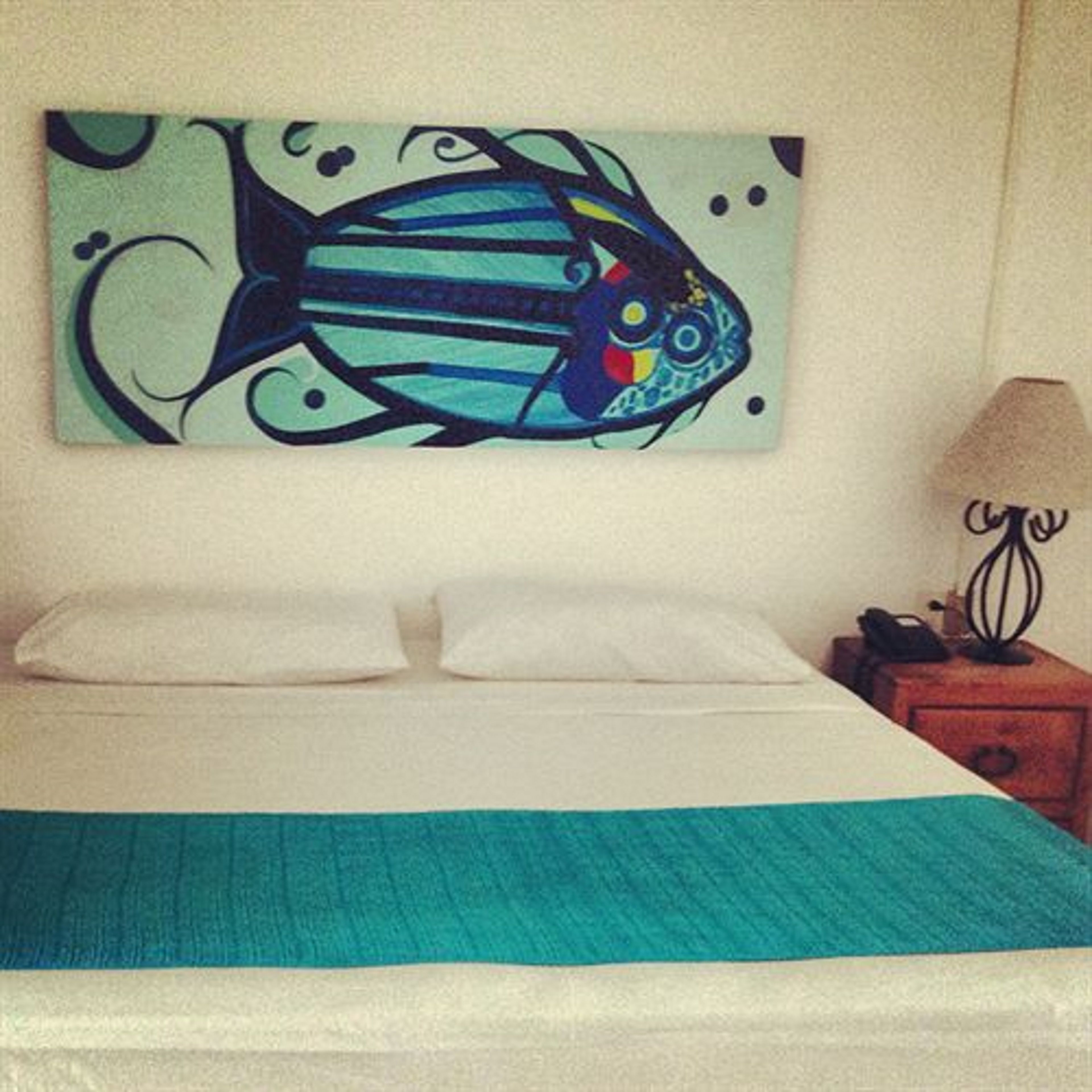 A bed with a teal bedspread, a white pillow, and a wall-mounted painting of a stylized fish.
