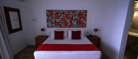 Habitación de hotel con una cama grande, una colcha roja y blanca, dos almohadas rojas, una obra de arte abstracta en la pared, una mesita de noche de madera con una lámpara, y un baño visible a través de una puerta abierta.