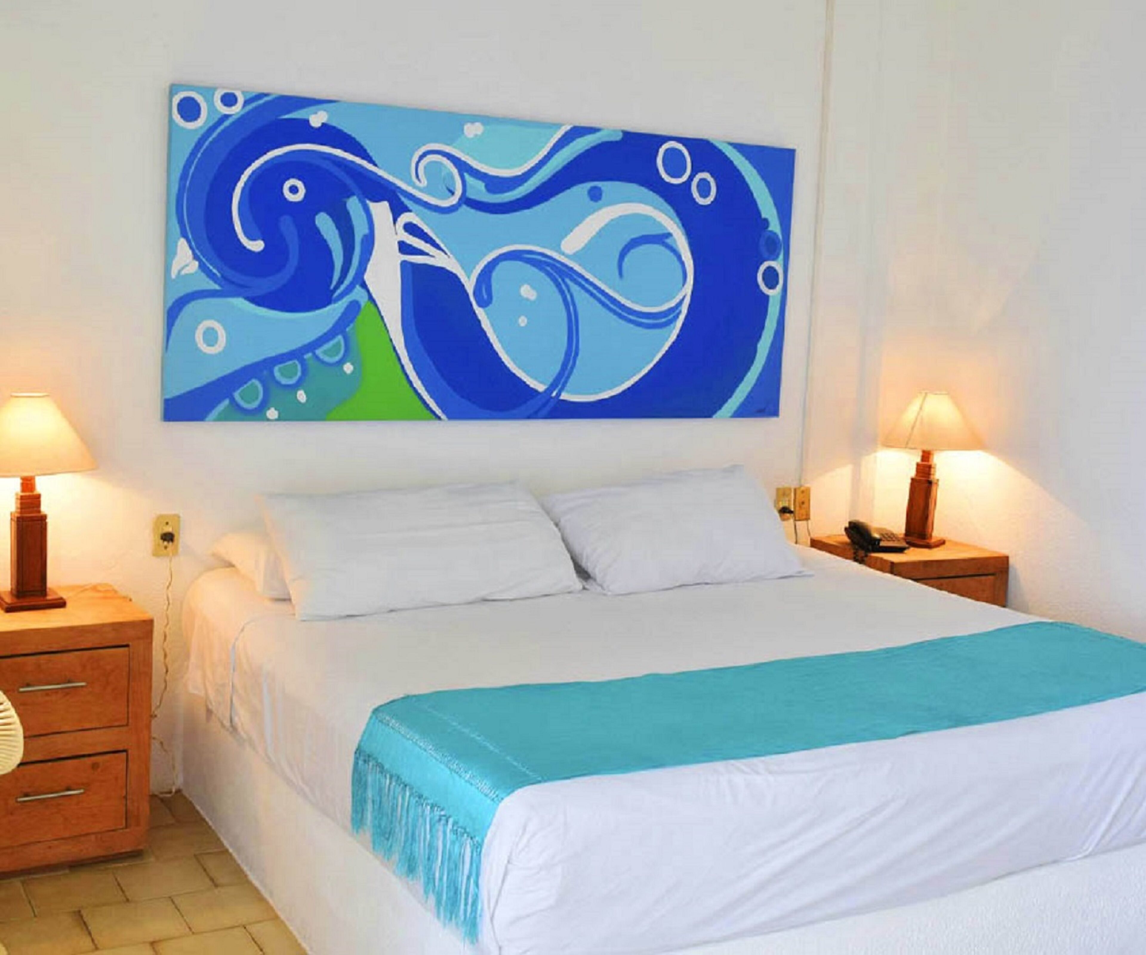A hotel room with a large bed, white bedding, a blue and white abstract painting on the wall, and two bedside lamps.