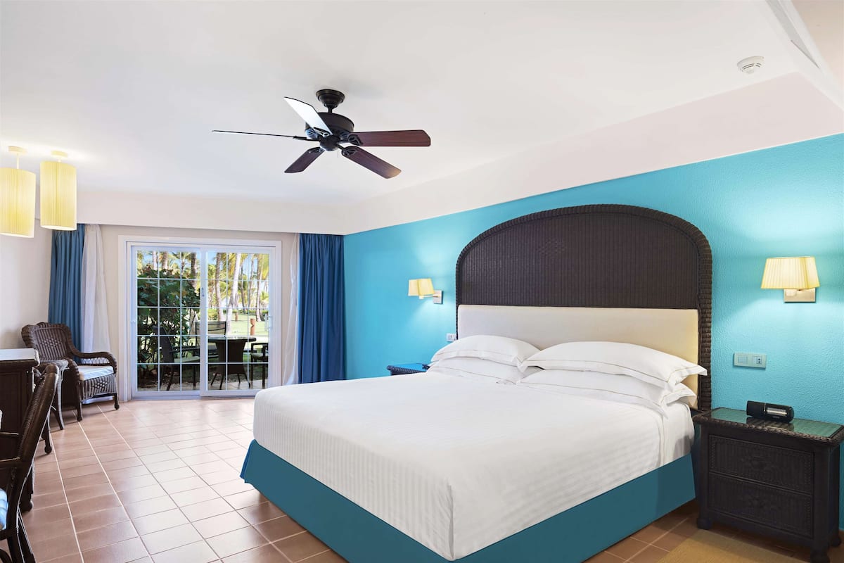 A hotel room with a bed, a ceiling fan, a nightstand, a lamp, a chair, and a view of the outdoors through a sliding door.