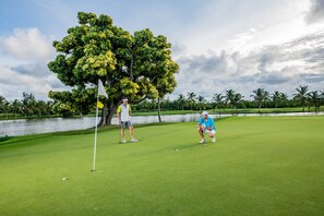 A golf course with a water body in the background, a large tree, and a clear sky.