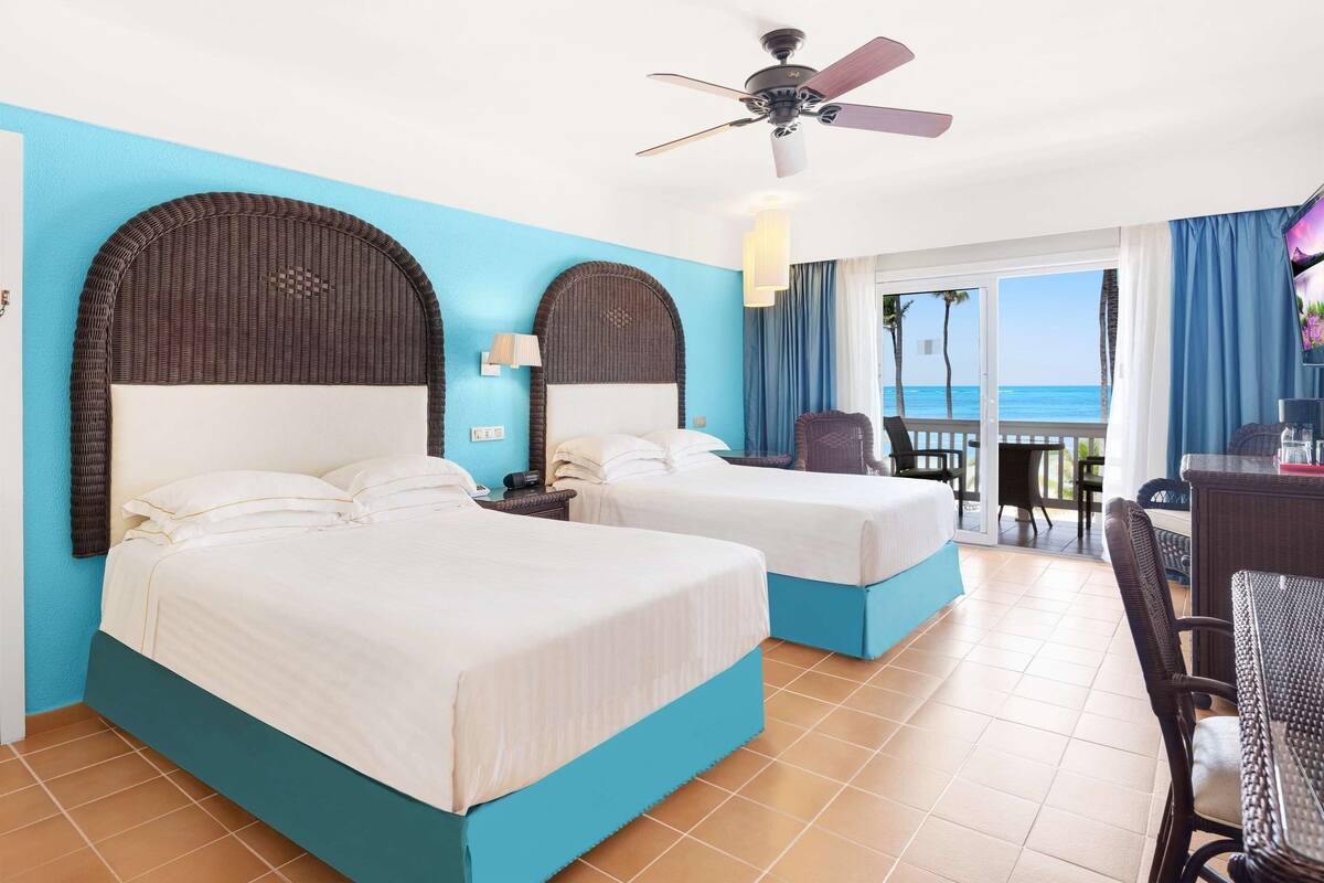 A hotel room with two beds, a ceiling fan, a desk with a chair, and a view of the ocean.