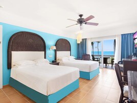 Habitación de hotel con dos camas, ventilador de techo, escritorio con silla y vista al océano.