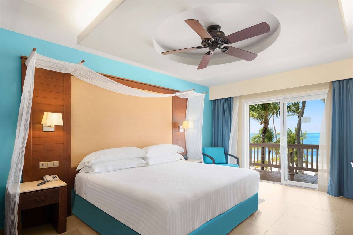 A hotel room with a bed, a nightstand, a desk, a chair, a ceiling fan, and a view of the ocean.