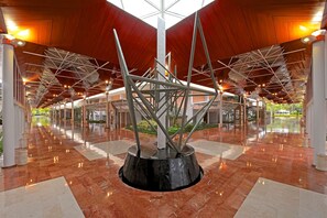 A spacious lobby with a modern geometric sculpture at the center, surrounded by polished marble floors and a high ceiling with a wooden finish.