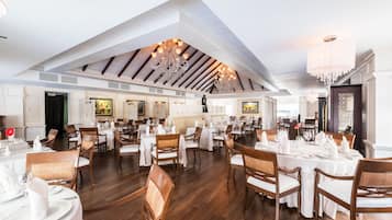 Un restaurant spacieux avec de hauts plafonds, des poutres en bois et des lustres. Les tables sont dressées avec des nappes blanches et des chaises.