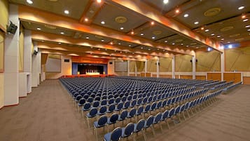 Een ruime zaal met rijen blauwe stoelen, een podium en een hoog plafond met inbouwverlichting.