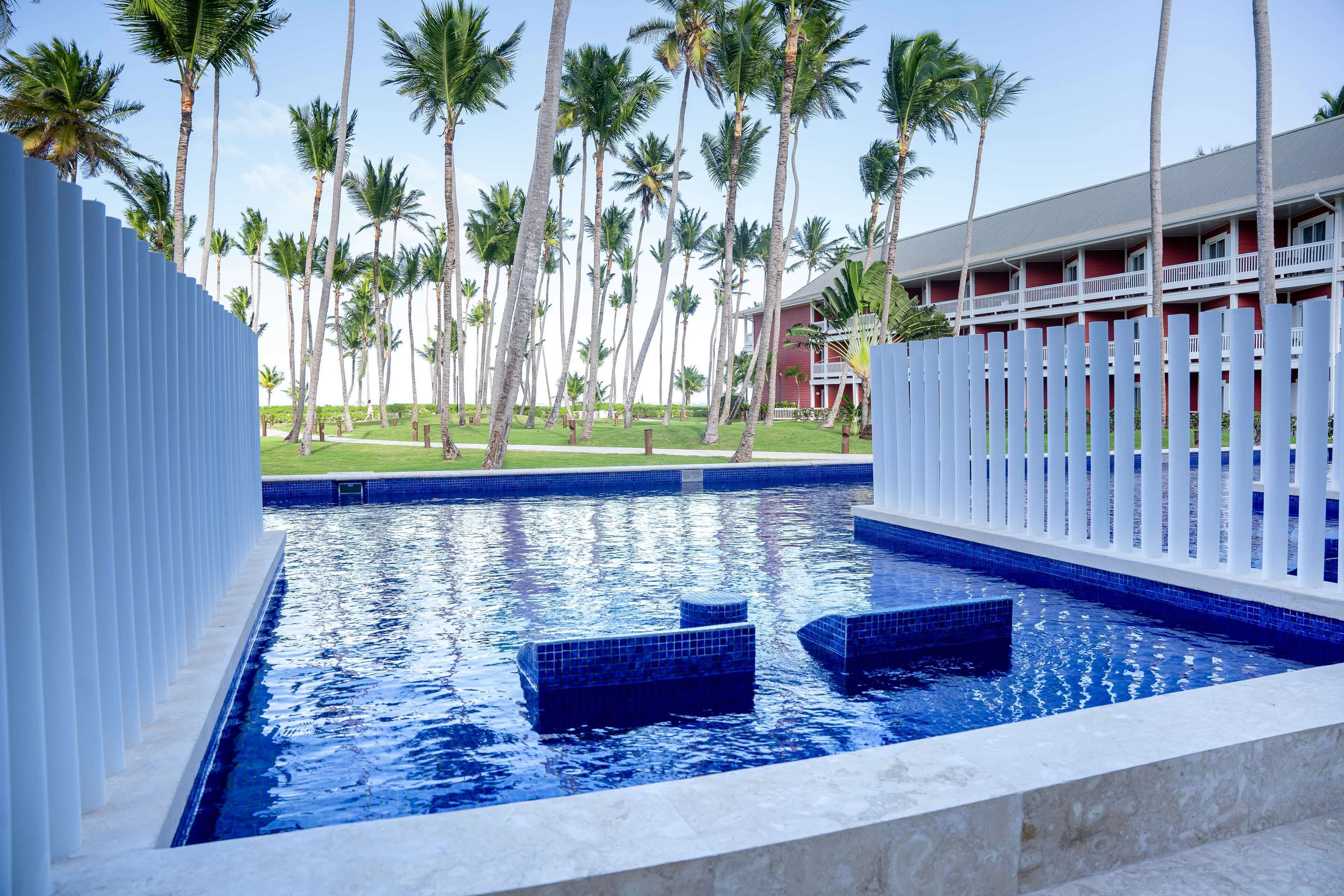 A swimming pool with blue tiles, surrounded by palm trees and a building with a red facade.