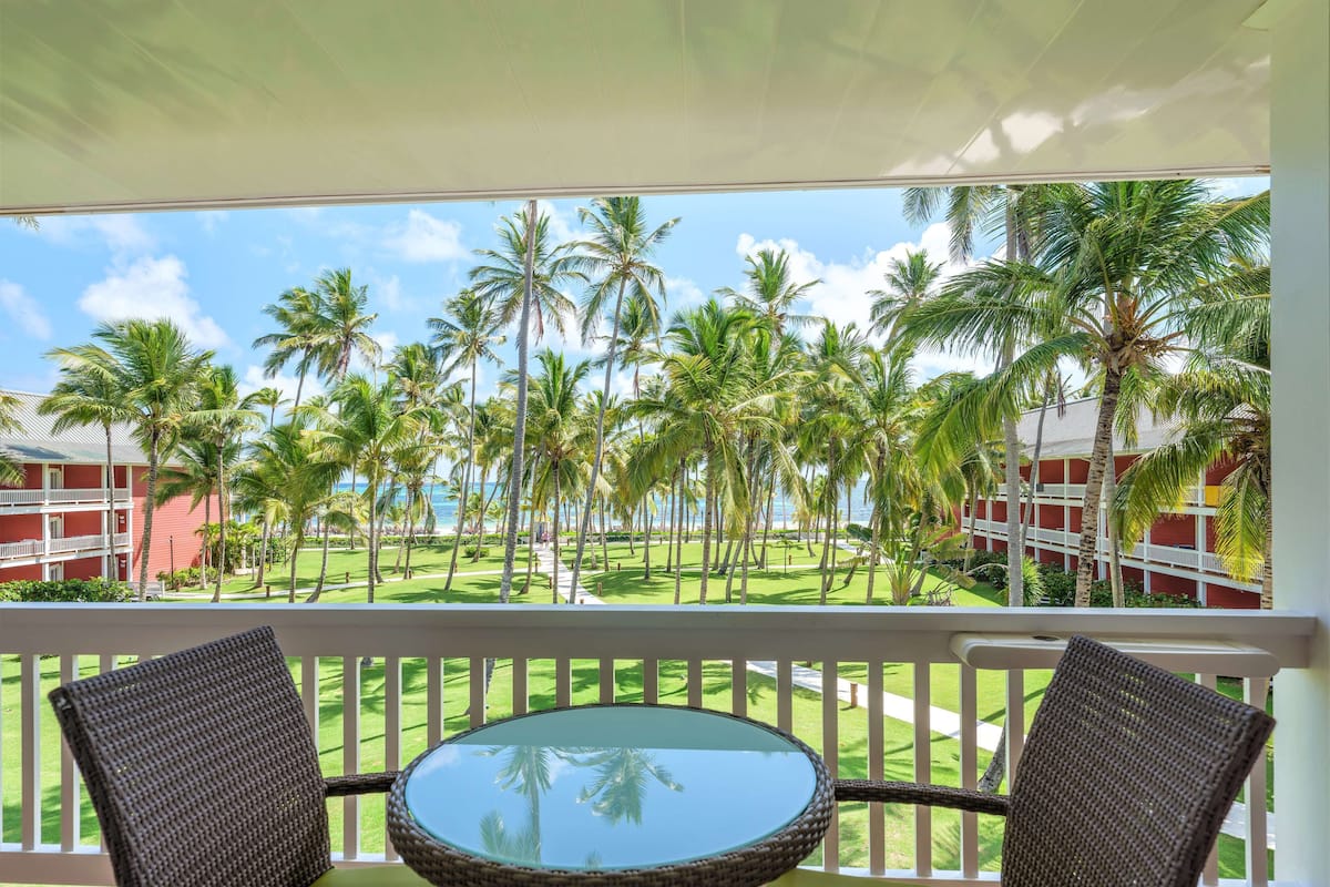 A balcony with wicker chairs and a glass table overlooking a lawn with palm trees and a building with balconies.