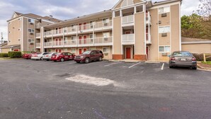 Un motel à plusieurs étages doté d’un parking, avec une entrée principale marquée par une porte rouge, de nombreux balcons et un espace de stationnement où sont garées plusieurs voitures.