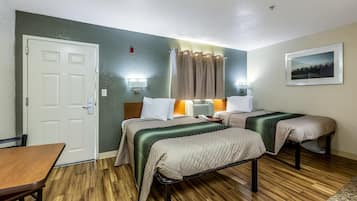 A hotel room with two beds, a wooden floor, a white door, a table, and a wall-mounted picture.