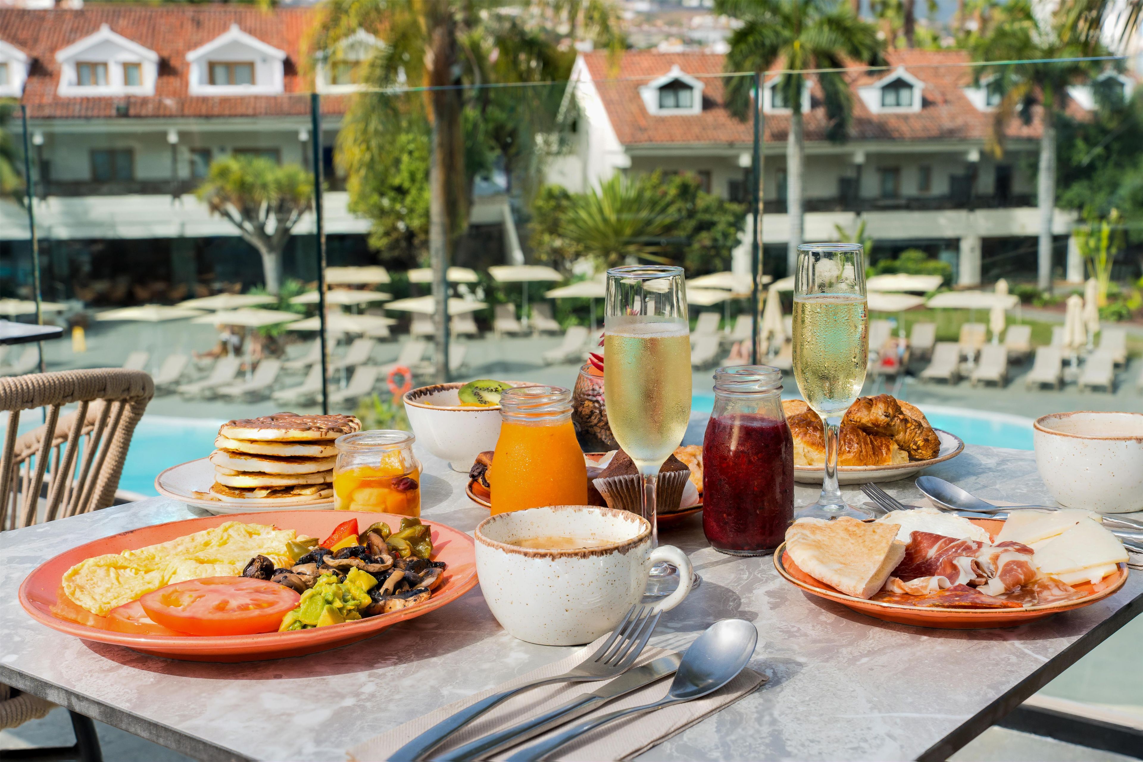 A breakfast table with various food items and drinks, overlooking a pool area with lounge chairs and umbrellas.