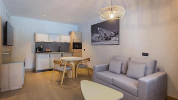Un espacio moderno para vivir con un sofá gris, una mesita blanca y una mesa de comedor con sillas. Hay una cocina con armarios y un microondas. Un cuadro de un paisaje montañoso cuelga en la pared.