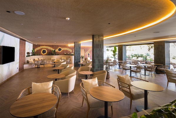 A spacious indoor area with multiple seating arrangements, including round tables and wicker chairs, and a large flat-screen TV mounted on the wall.