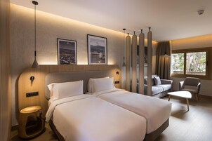 Habitación de hotel moderna con dos camas, un sofá, una silla y una mesita. Hay cuadros enmarcados en la pared, una ventana con cortinas y una lámpara de techo.