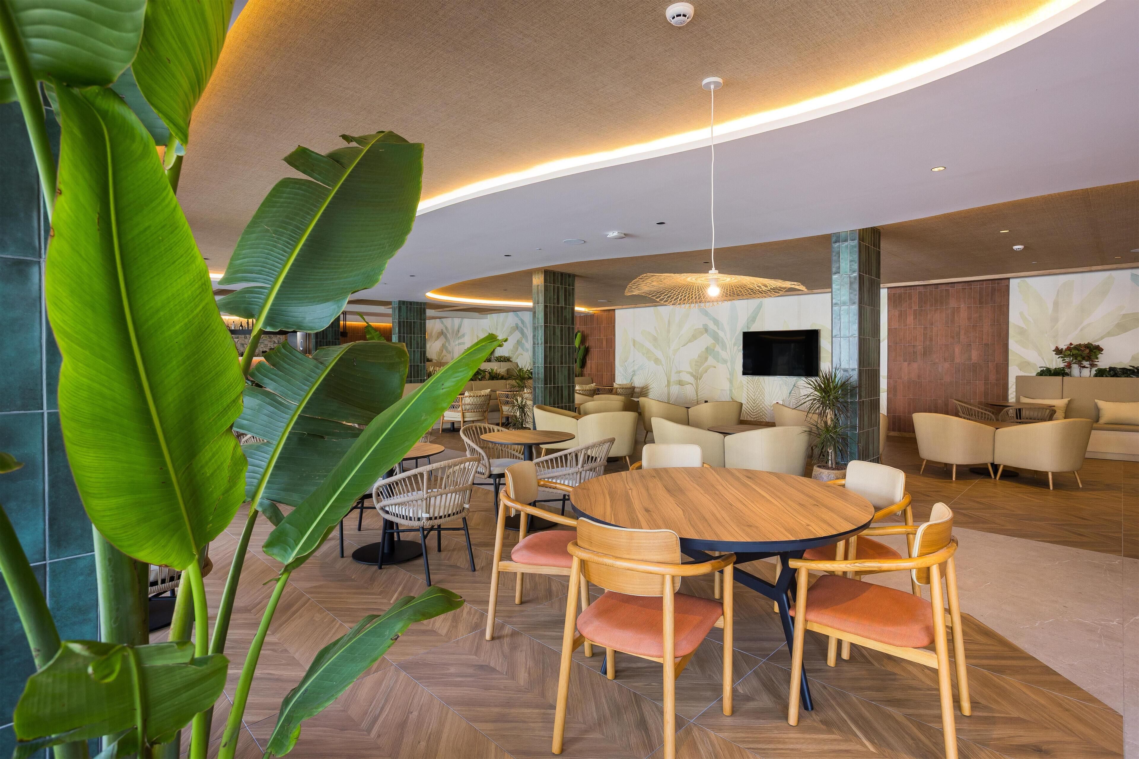 Een moderne ruimte met een ronde houten tafel, omringd door stoelen, een grote hanglamp en diverse zithoeken met banken en stoelen.