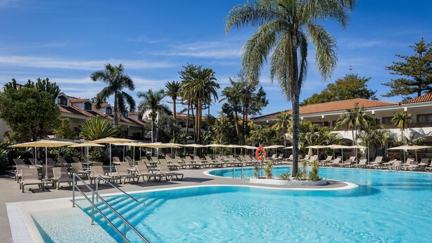 A pool area with lounge chairs and umbrellas, surrounded by palm trees and buildings. - Radisson Resort & Residences Tenerife - Adults Only (Puerto de la Cruz)