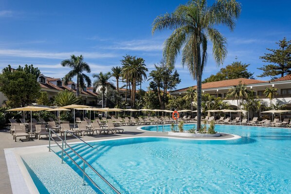 Zona de piscina con sillas de descanso y sombrillas, rodeada de palmeras y edificios.