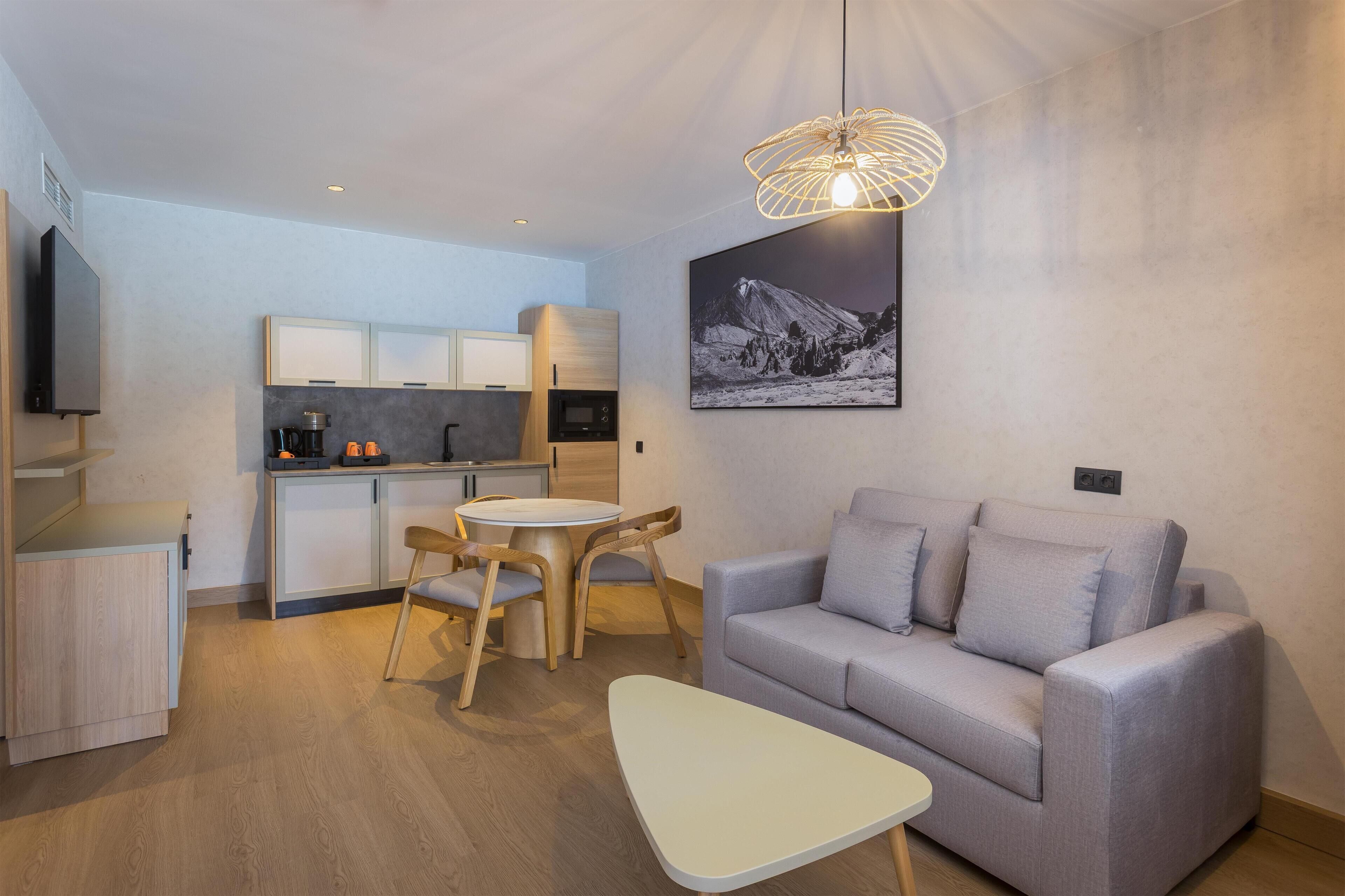 Un espacio habitable moderno con cocina, un sofá gris, una mesita blanca pequeña y una zona de comedor con sillas de madera.