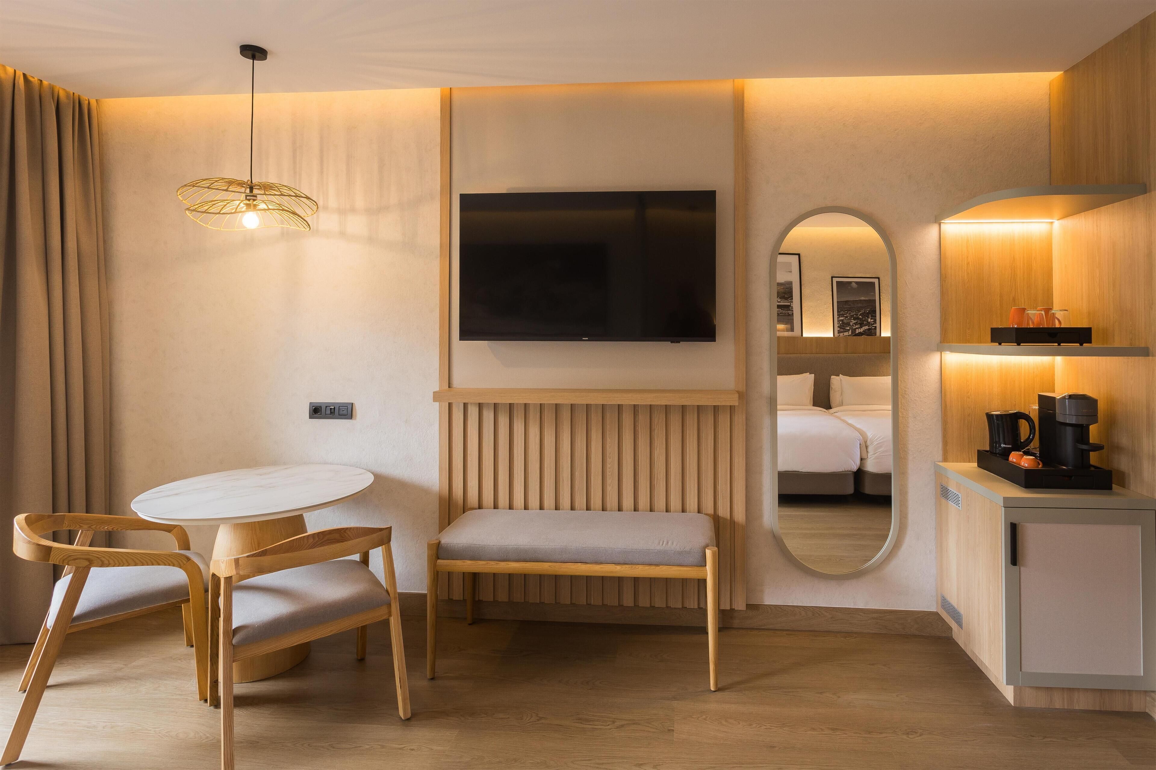 Habitación de hotel con una mesa redonda, dos sillas de madera, un banco, un televisor montado en la pared, un espejo, un mueble pequeño con una cafetera y piso de madera.
