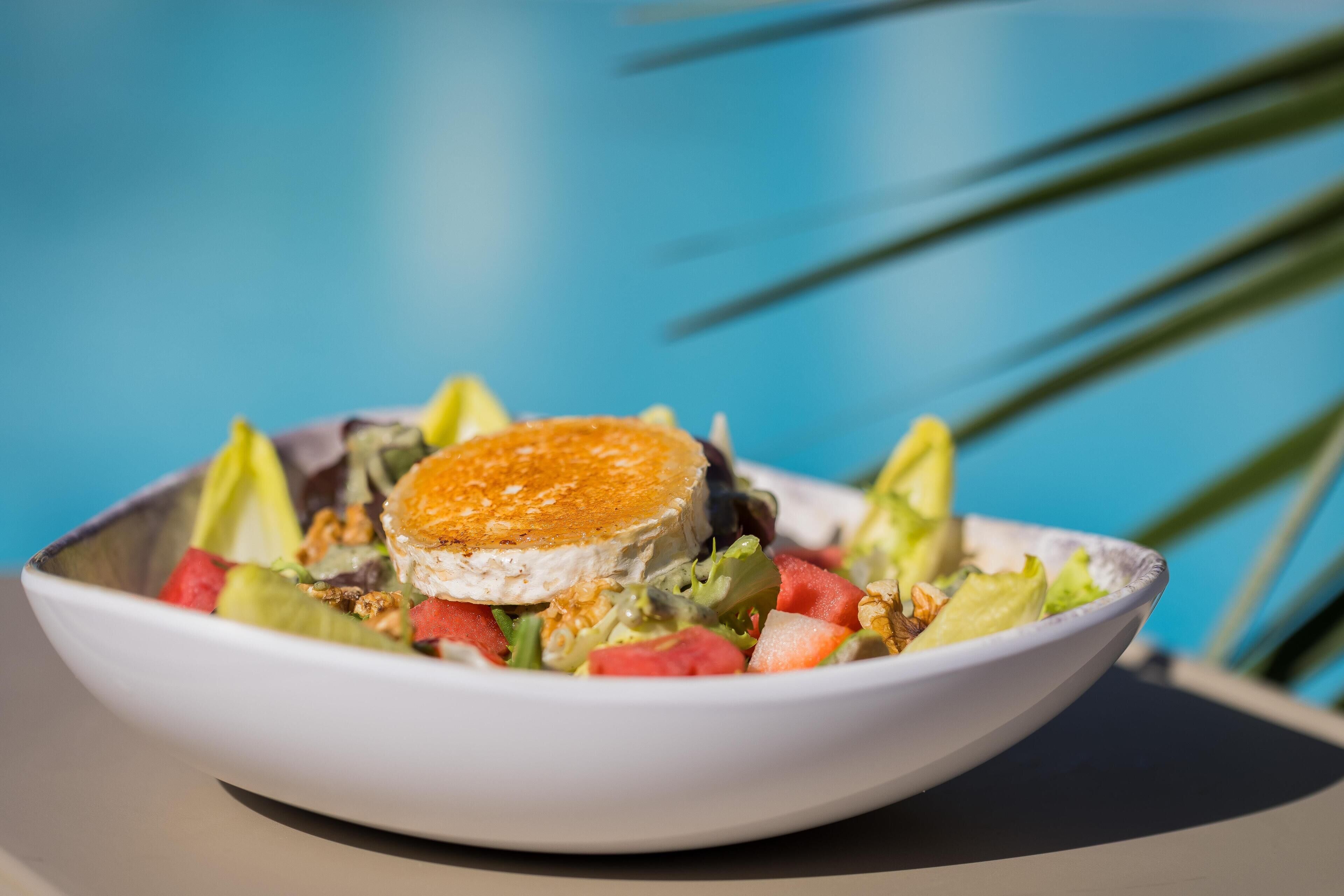 Un bol de salade surmonté d’une tranche de fromage, posé sur une table devant un fond bleu.