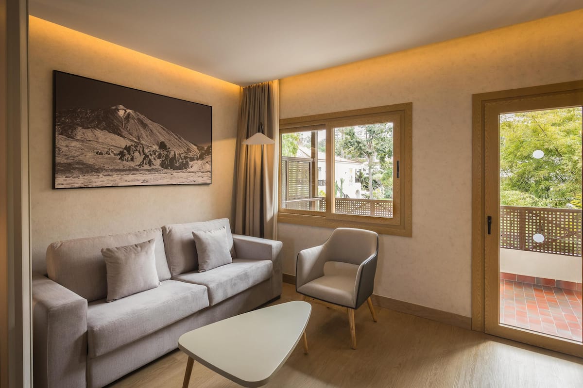 Una sala moderna con sofá, sillón y una mesa de centro. Hay un cuadro grande en la pared y una ventana con vistas a zonas verdes.