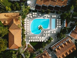 Vista aérea de un complejo turístico con una piscina grande, varias zonas de descanso y palmeras alrededor.