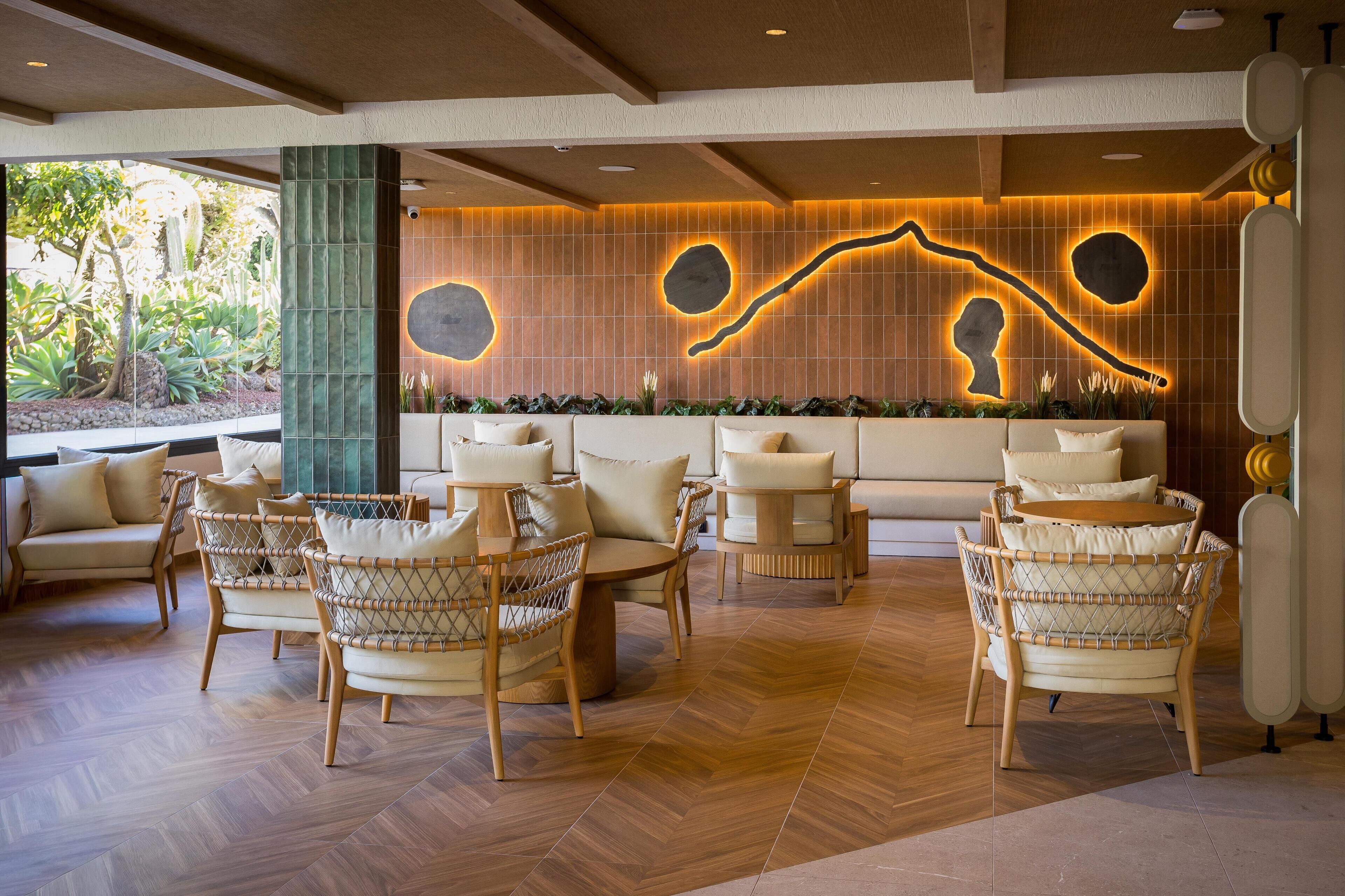 A modern lounge area with wicker furniture, a large wall with illuminated designs, and a view of greenery outside.
