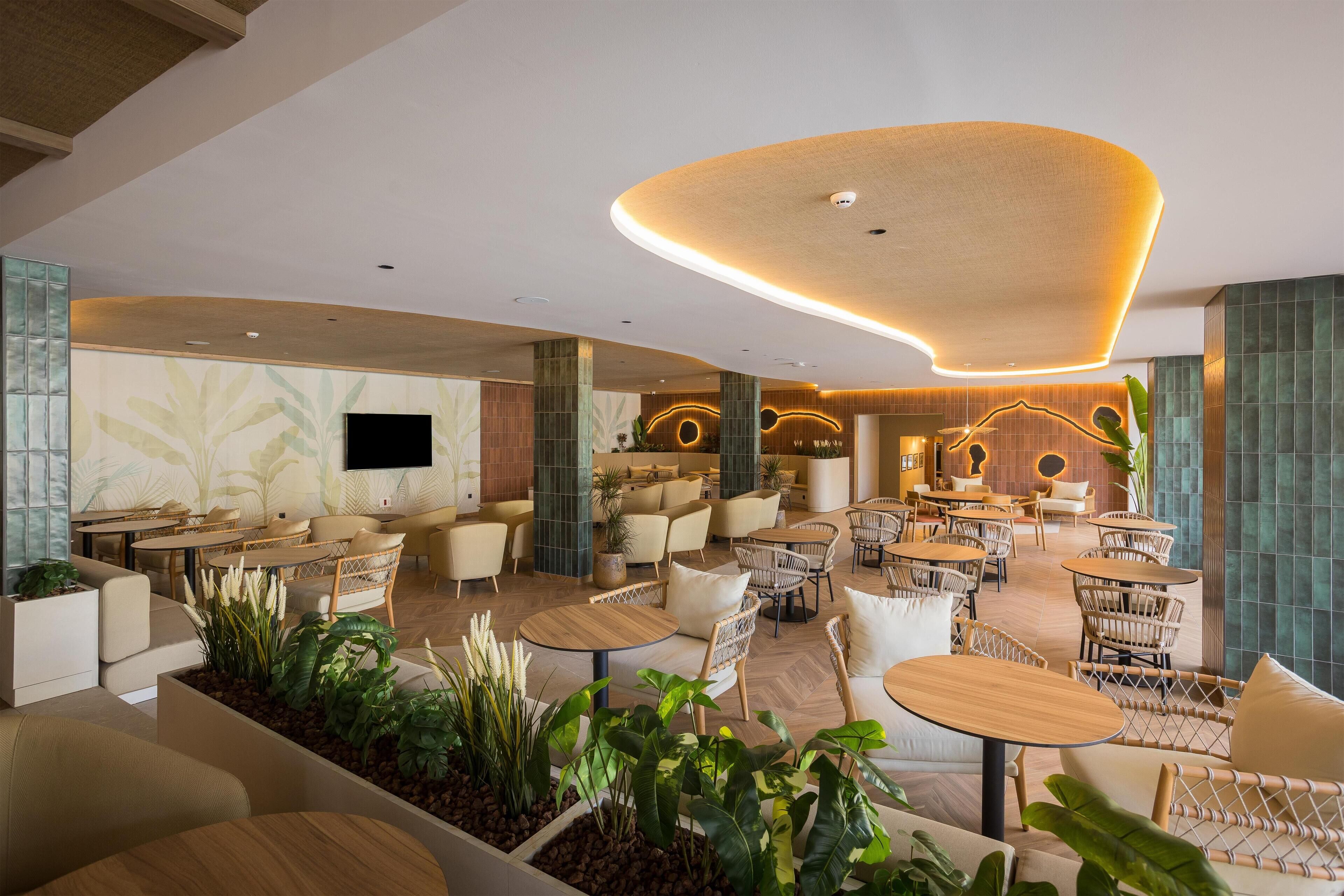 Un vaste espace intérieur aménagé avec différents types de sièges, notamment des tables rondes et des fauteuils confortables, le tout agrémenté de plantes en pot.