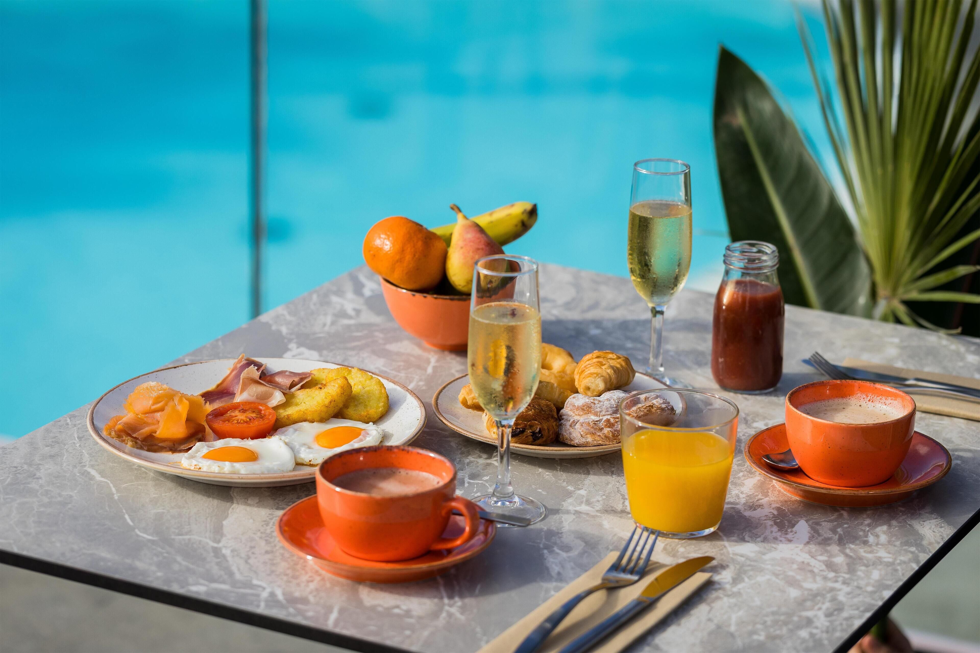 Una mesa de desayuno con platos de comida, bebidas y vista a una piscina.