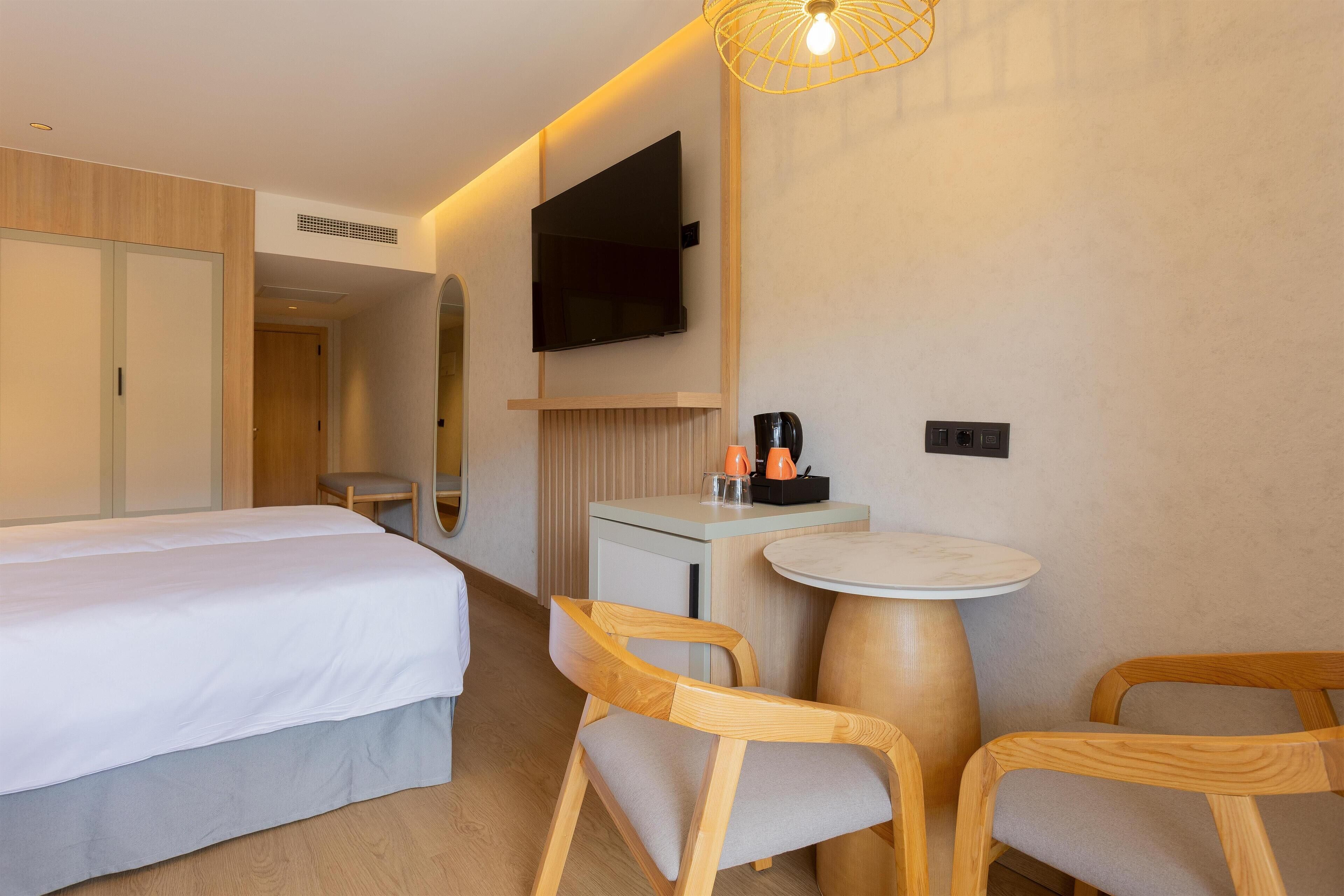 Habitación de hotel con cama, televisión montada en la pared, una mesita con sillas y cafetera.
