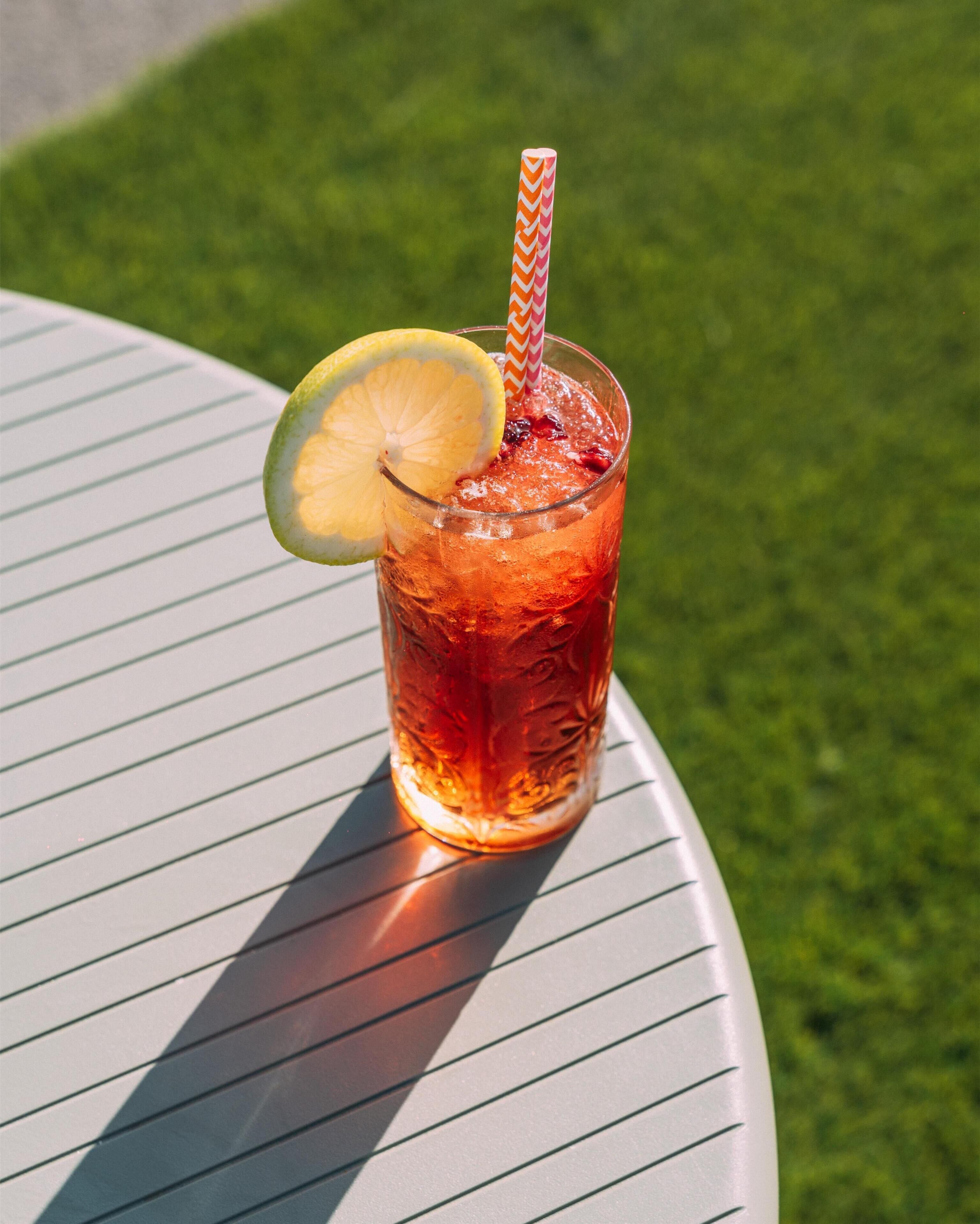 A glass of iced tea with a lemon slice and a striped straw on a white table with green grass in the background.