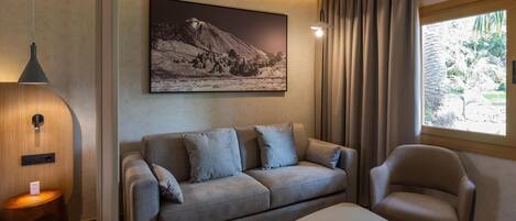 Ein modernes Wohnzimmer mit Sofa, Sessel und Tisch. Ein großes gerahmtes Foto hängt an der Wand, und es gibt ein Fenster mit Vorhängen.