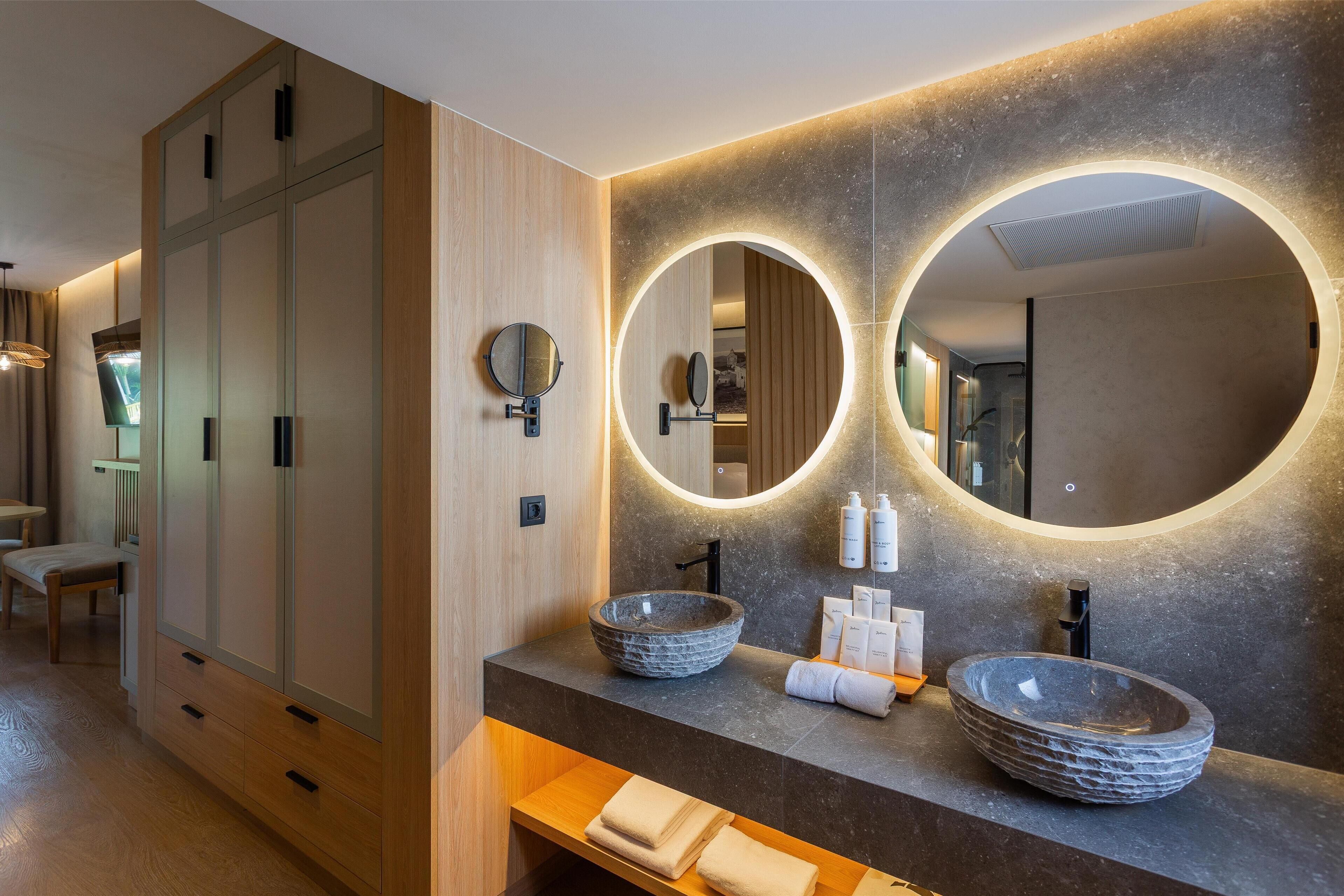 A modern bathroom with a wooden vanity, two vessel sinks, and a large mirror with circular lighting.