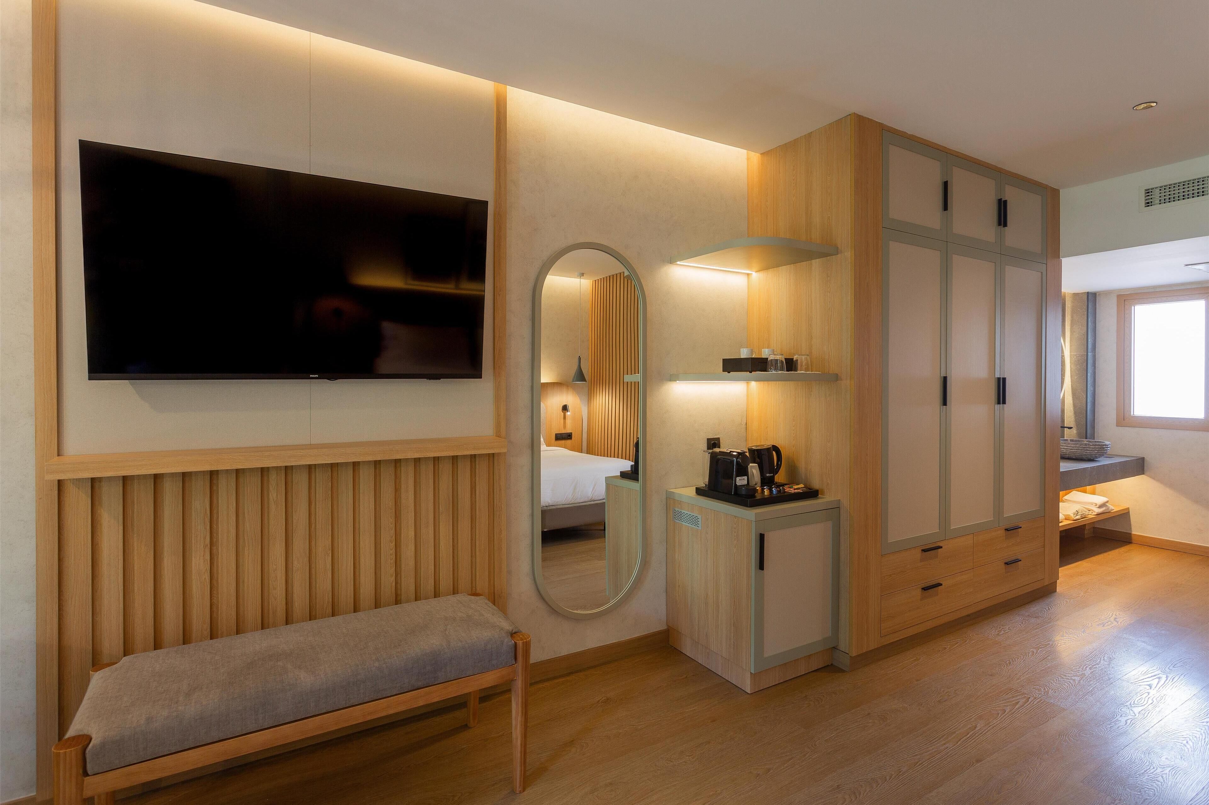 A hotel room with a wooden bench, a TV mounted on the wall, a mirror, a wardrobe, and a bedside table with a lamp.