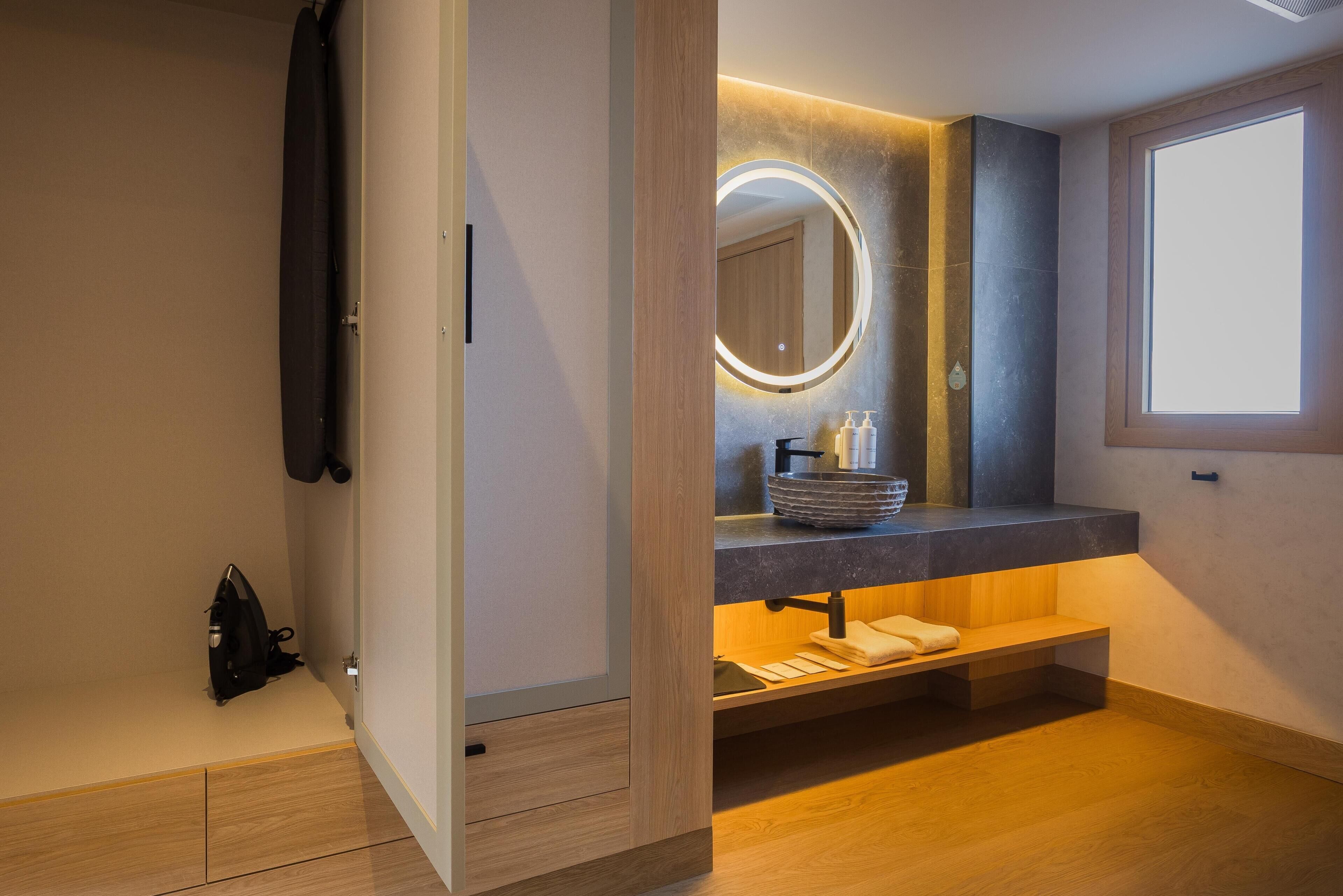 A modern bathroom with a wooden vanity, a round mirror with lighting, a sink with a soap dispenser, and a window providing natural light.