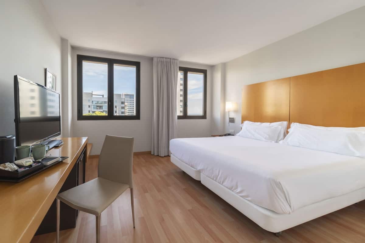 Habitación de hotel con cama, escritorio con televisor, silla y ventanas amplias que ofrecen vistas a la ciudad.
