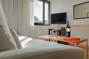 A modern hotel room with a sofa, a small table with a bottle of wine and two glasses, a book, a television, and a window with curtains. - Catalonia Barcelona Beach (Barcelona)