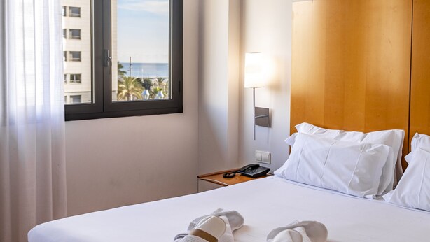 A hotel room with a large bed, white linens, a wooden headboard, a nightstand with a lamp, and a view of the sea through a window.